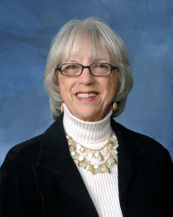 A smiling woman with glasses and short, gray hair is wearing a turtleneck and a decorative necklace against a blue background.