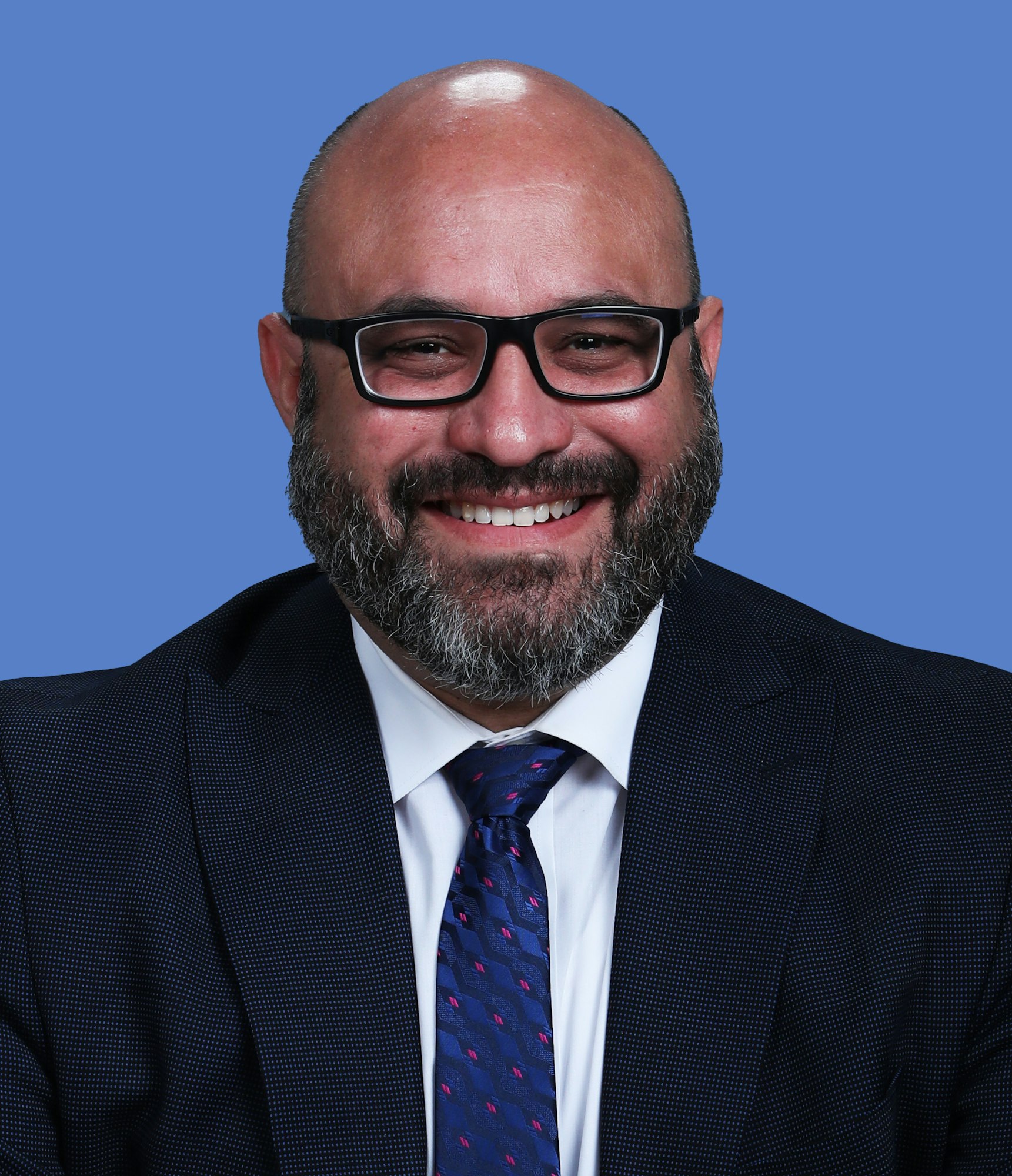 A smiling person with a beard, wearing glasses, a suit, and a tie, against a blue background.