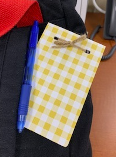 A blue pen next to a yellow checkered notepad with a twine loop, resting on fabric.