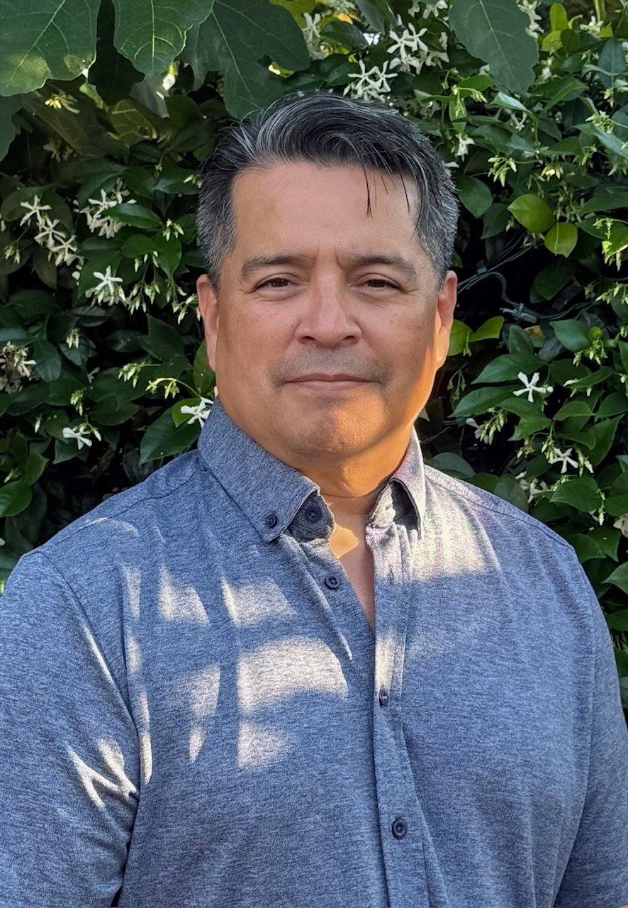 A man stands outdoors, smiling under natural light, with green foliage and small flowers in the background.