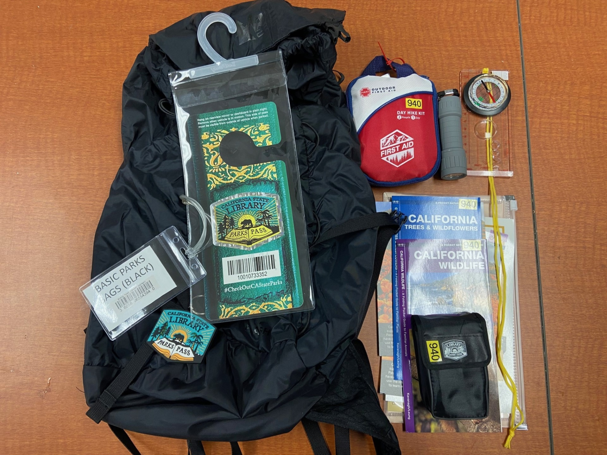 A backpack, parks pass, first aid kit, flashlight, compass, map case, wildlife guides, and a pouch on a table.