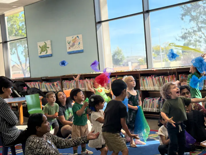 A lively scene in a library with children playing and laughing, some throwing colorful scarves, while adults observe nearby.