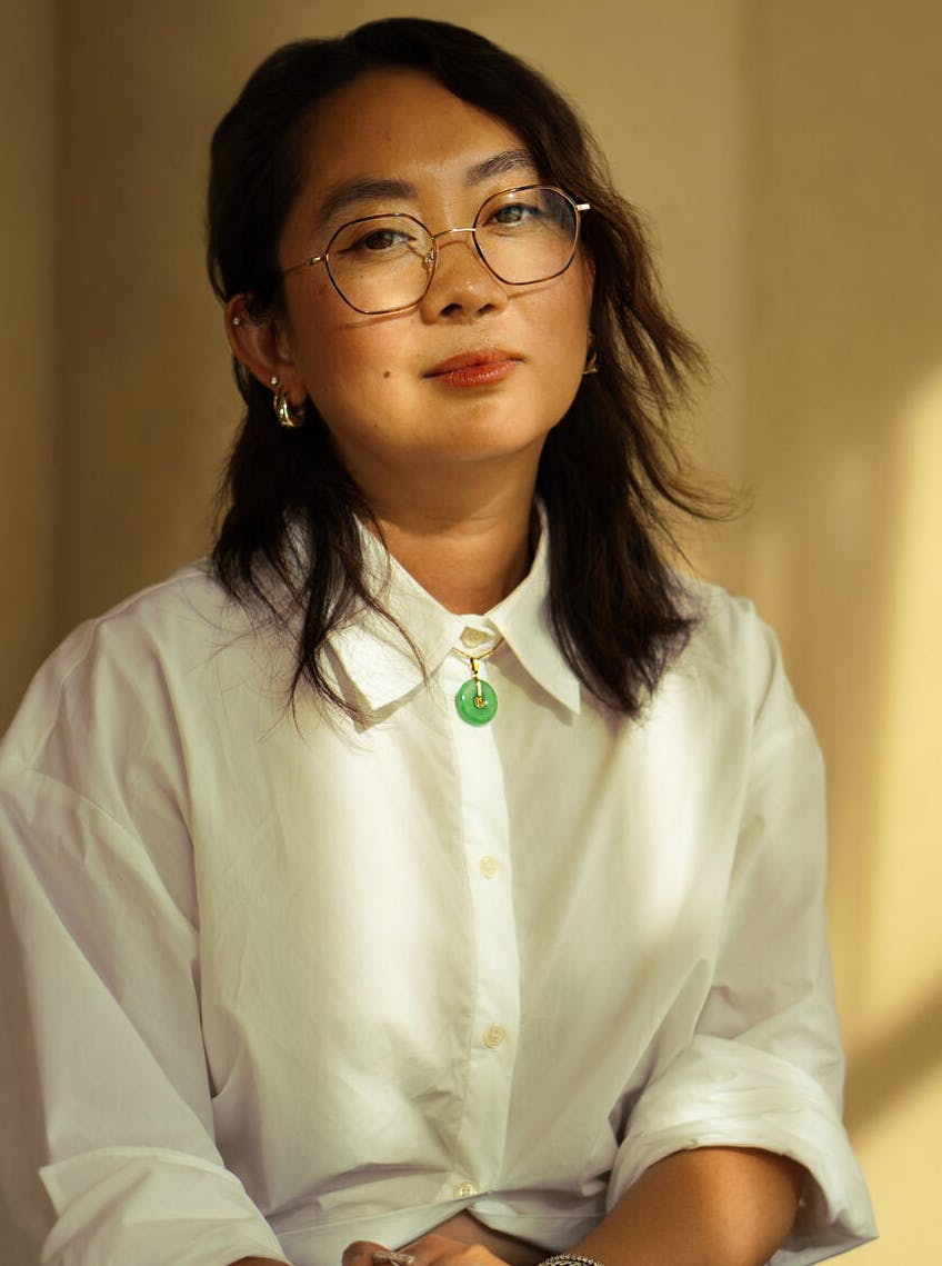 A person in glasses and a white shirt, sitting with hands folded, in soft lighting.