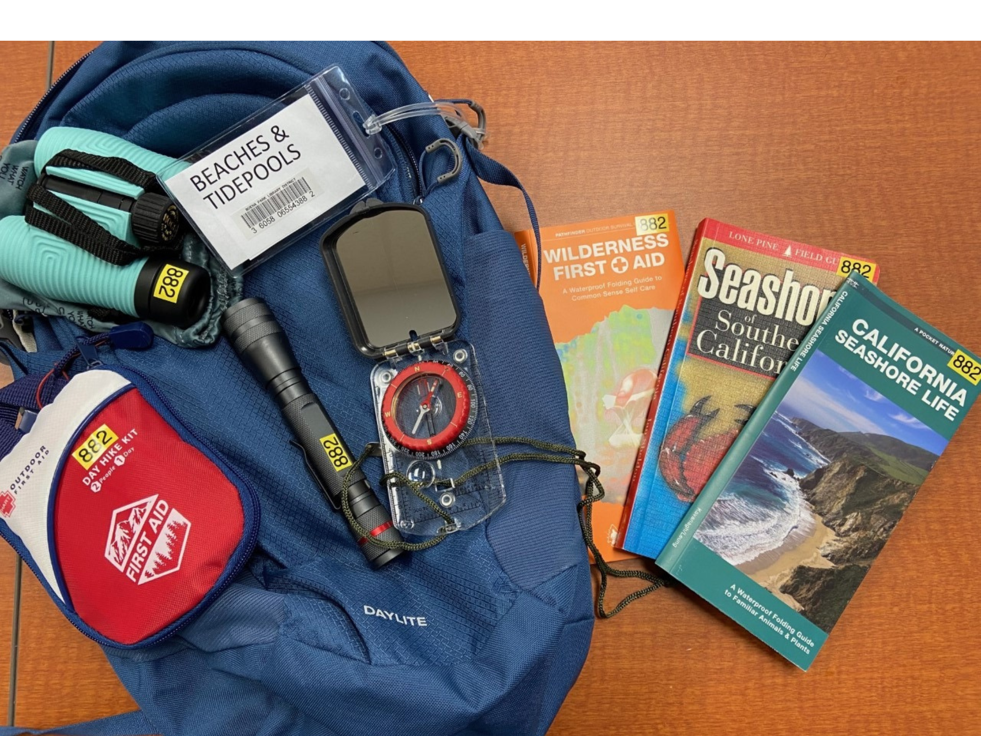 Outdoor gear with a backpack, flashlight, compass, first aid kit, books on wilderness, and tide pools guide.
