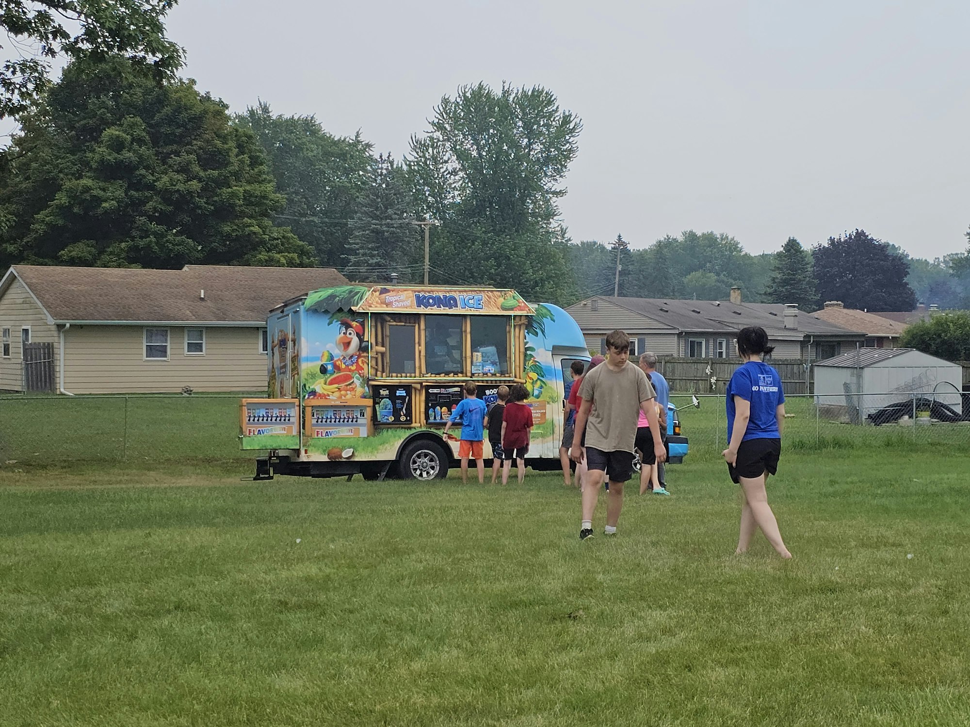 People gathered around a colorful Kona Ice truck in a grassy park setting.