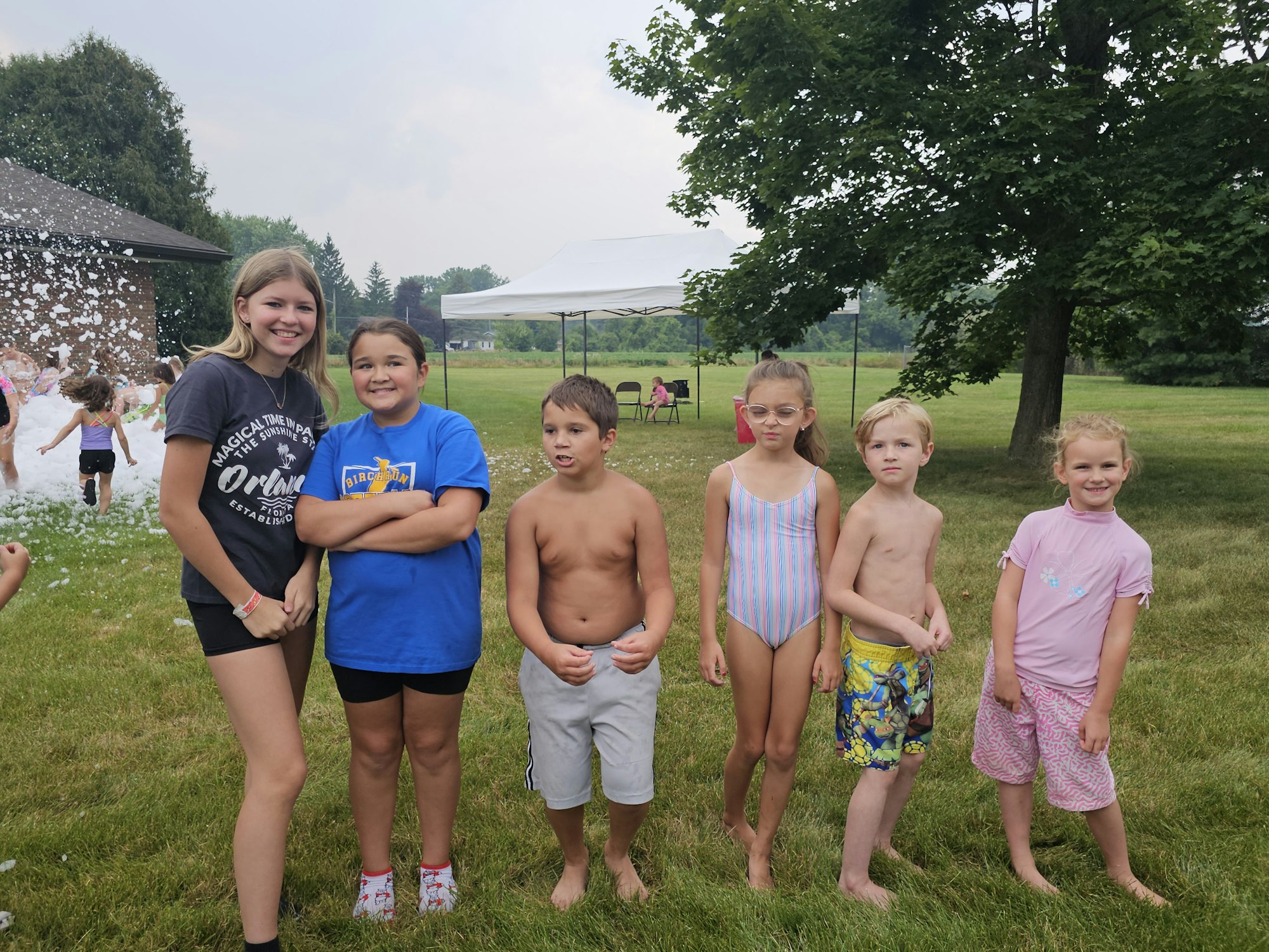 A group of kids in swimwear stand on grass, with a foam party in the background.