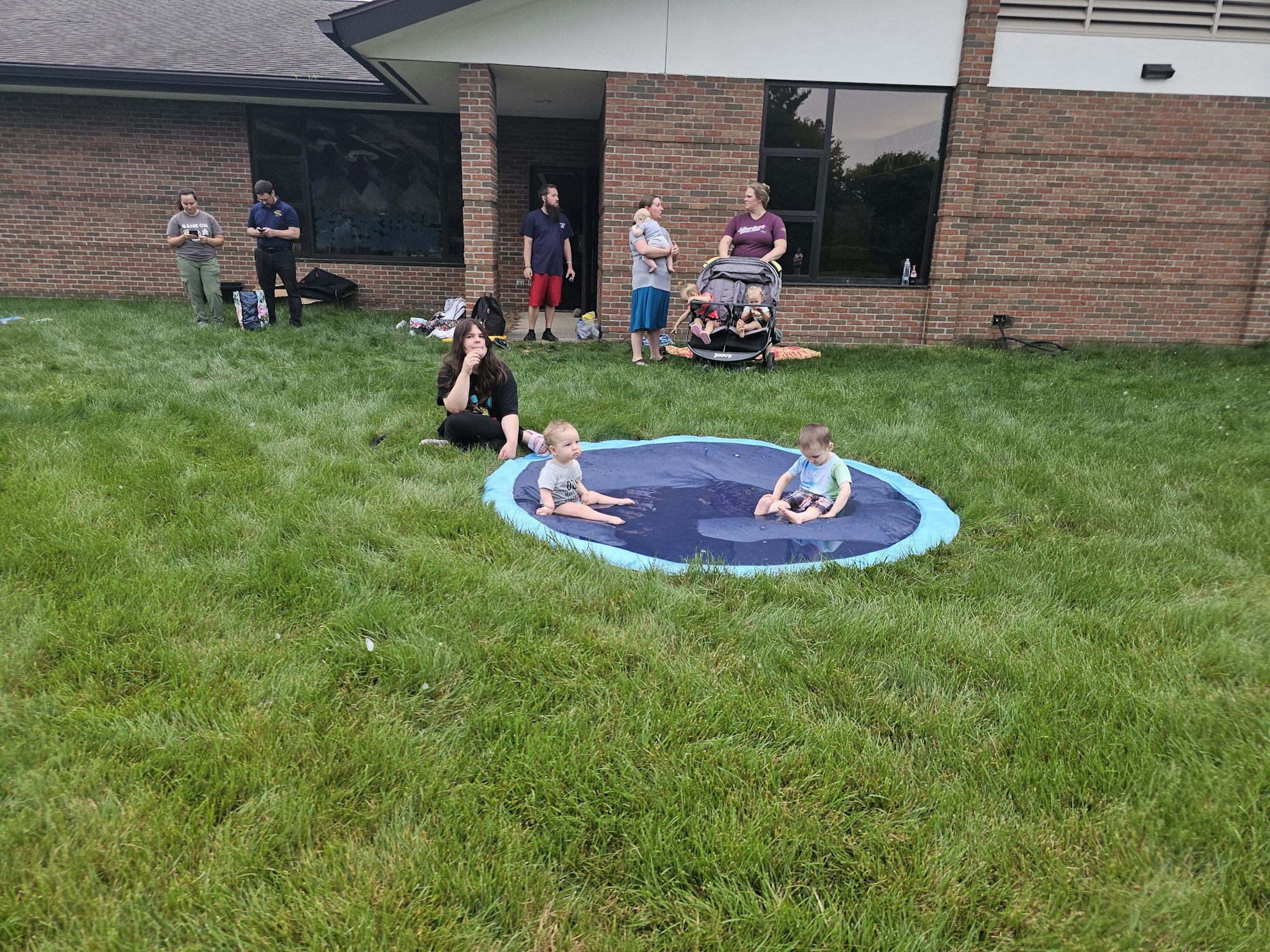 Children playing on a splash pad in a grassy yard, surrounded by adults taking photos.