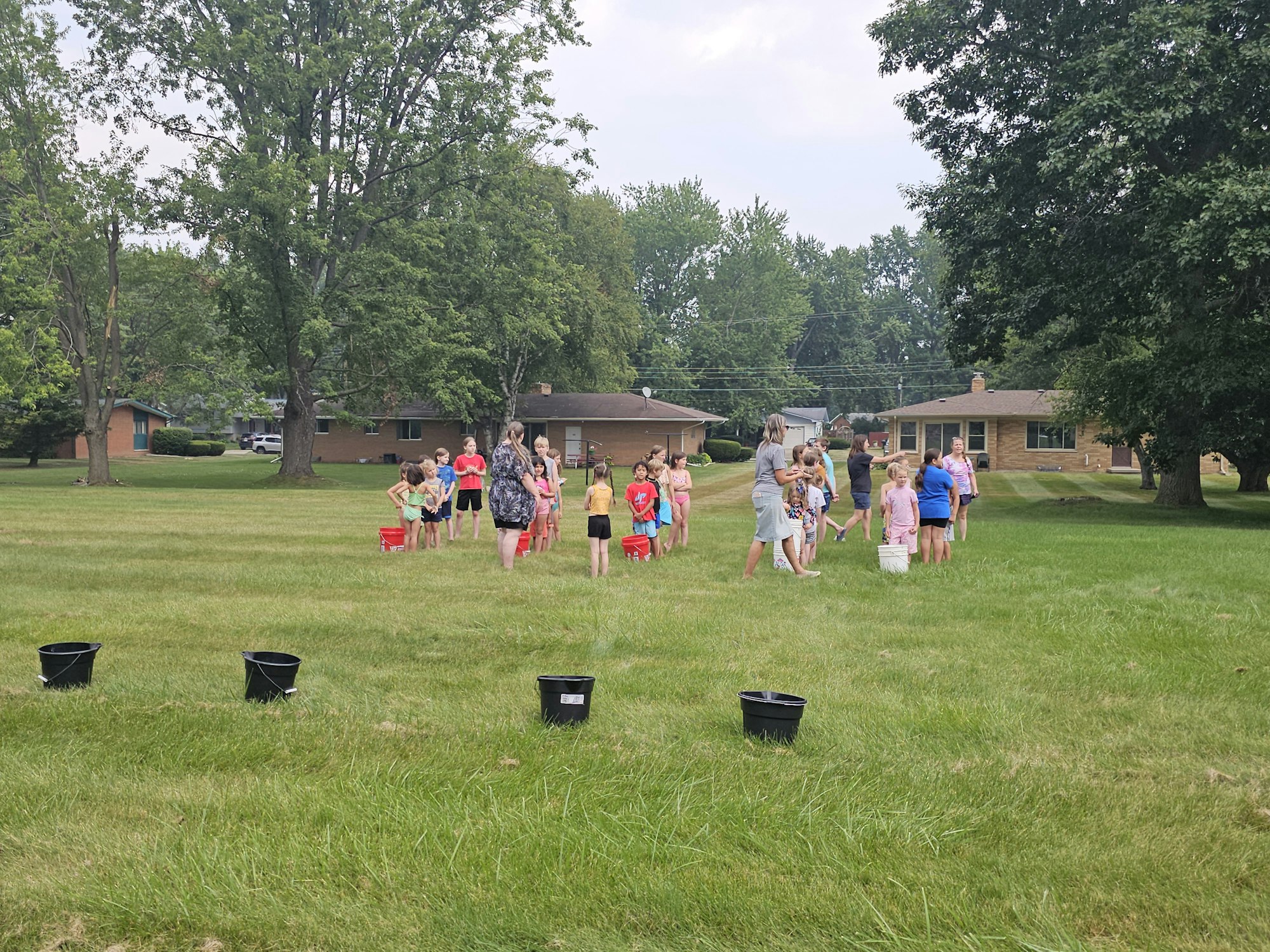 A group of children doing a relay race on a grassy field, with houses and trees in the background.