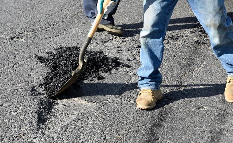 A person is repairing a road by using a shovel to apply black asphalt to a damaged area of pavement.