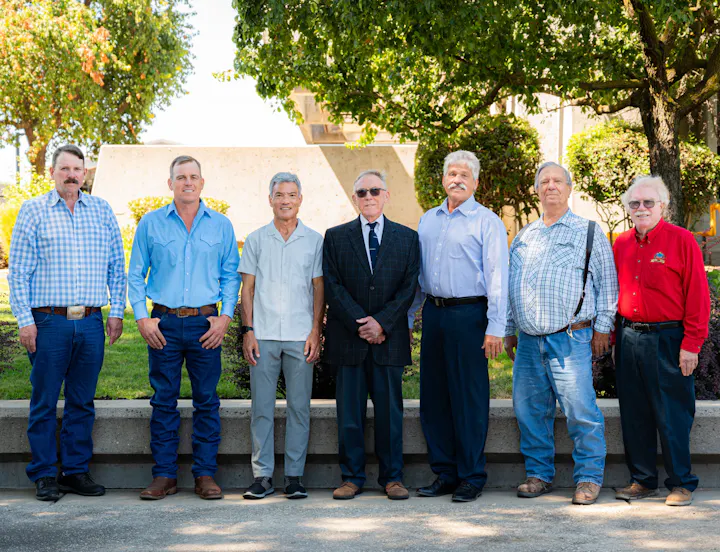 A group of seven men dressed in casual and formal attire poses outdoors in a sunny setting.