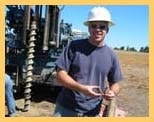 A person with a hard hat standing in front of drilling machinery in a field.