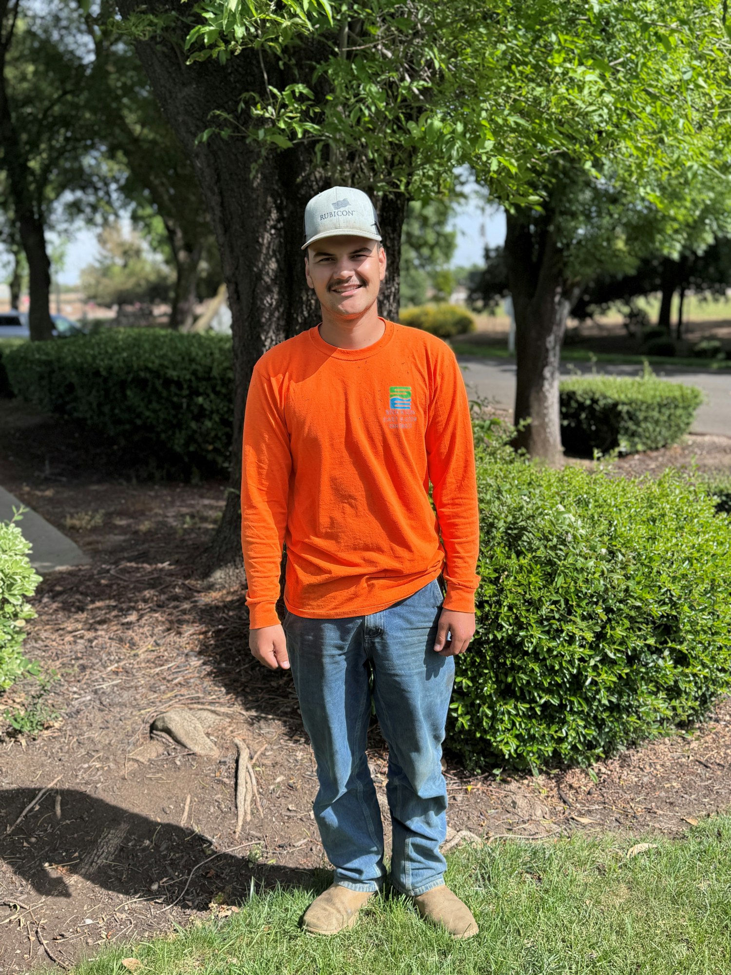A young man wearing an orange long-sleeve shirt, jeans, and a gray cap stands outdoors among green shrubs and trees.