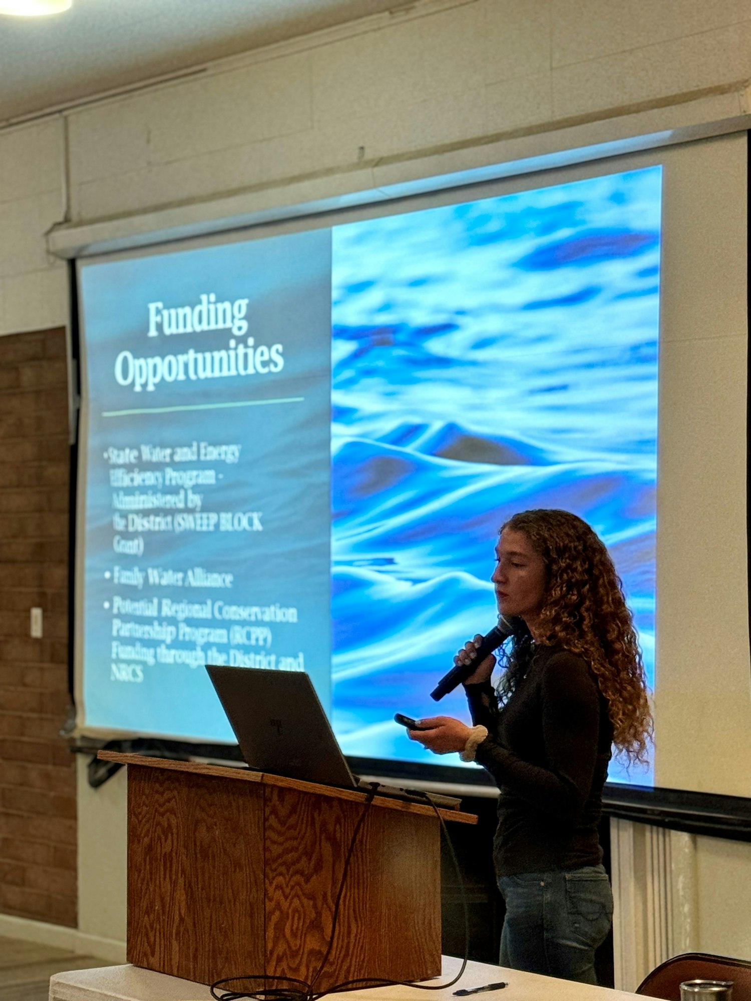 A person presenting with a microphone in front of a screen displaying "Funding Opportunities".