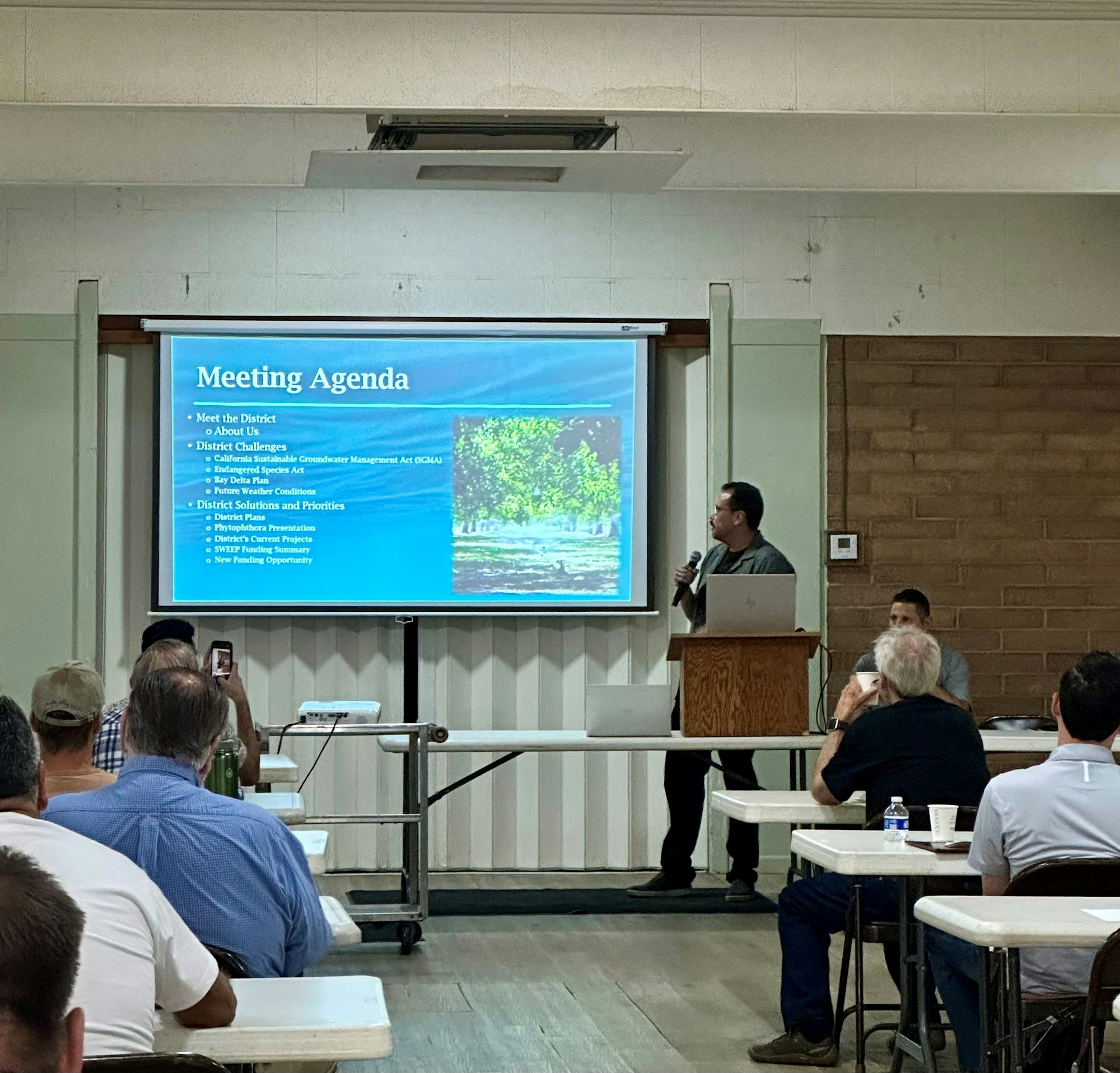 A man presents a meeting agenda on a projected screen in a room with seated attendees.