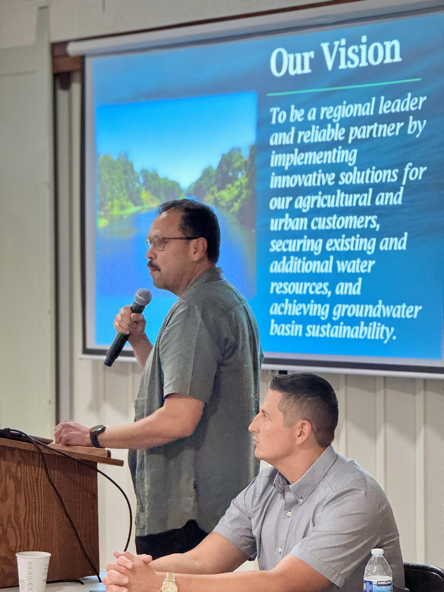 A man speaks at a podium with a vision statement about water management on a screen. Another person sits at a table.