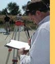 Person with clipboard and surveying equipment outdoors.