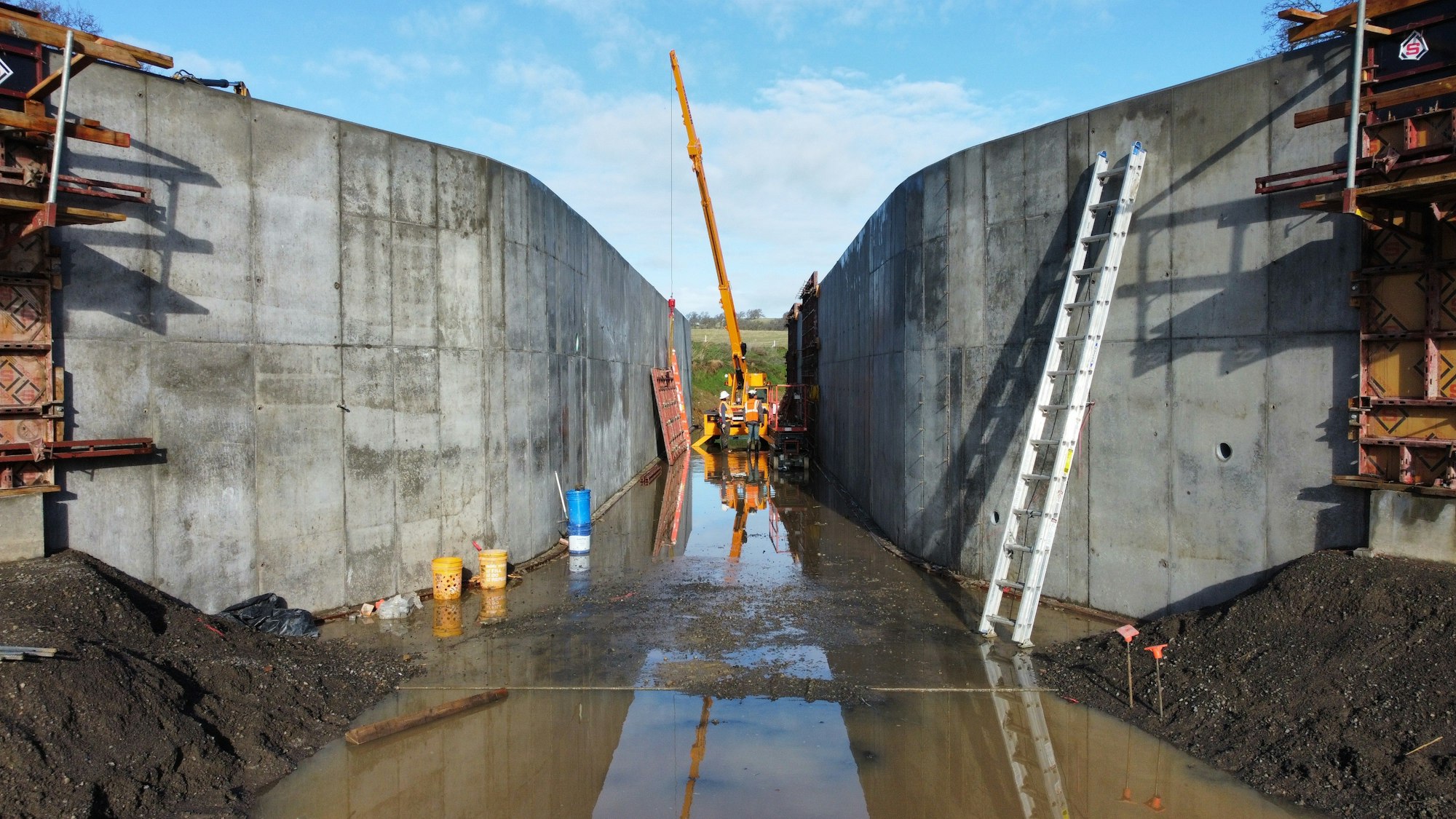 A construction site with a crane between concrete walls, ladders, and equipment, with water on the ground.