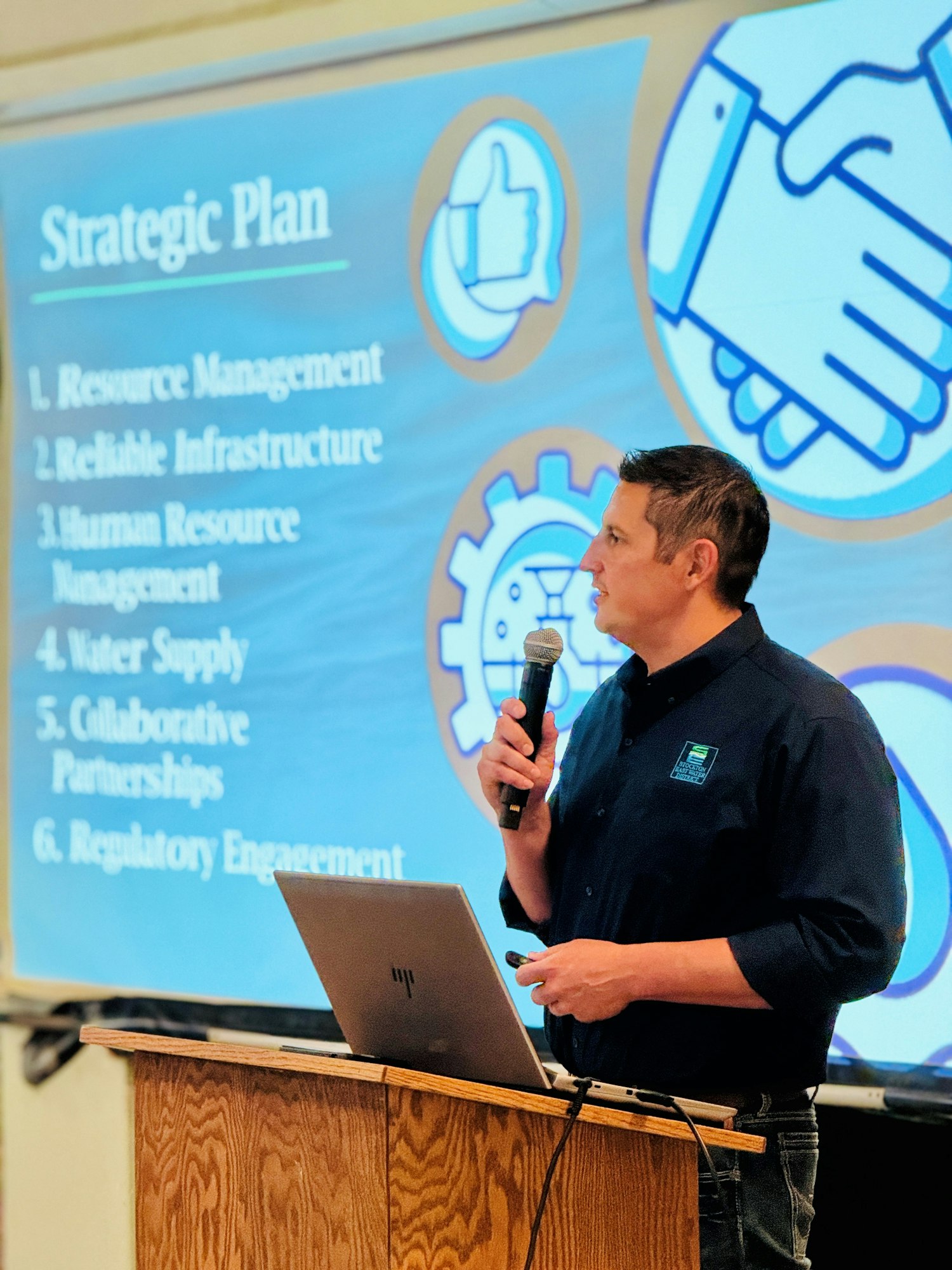 A person presenting a strategic plan with topics like resource management and infrastructure on a projector screen.
