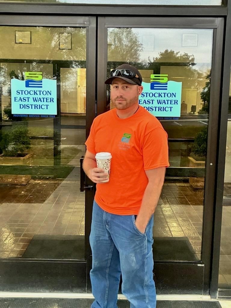 A person in an orange shirt stands outside the Stockton East Water District building, holding a coffee cup.