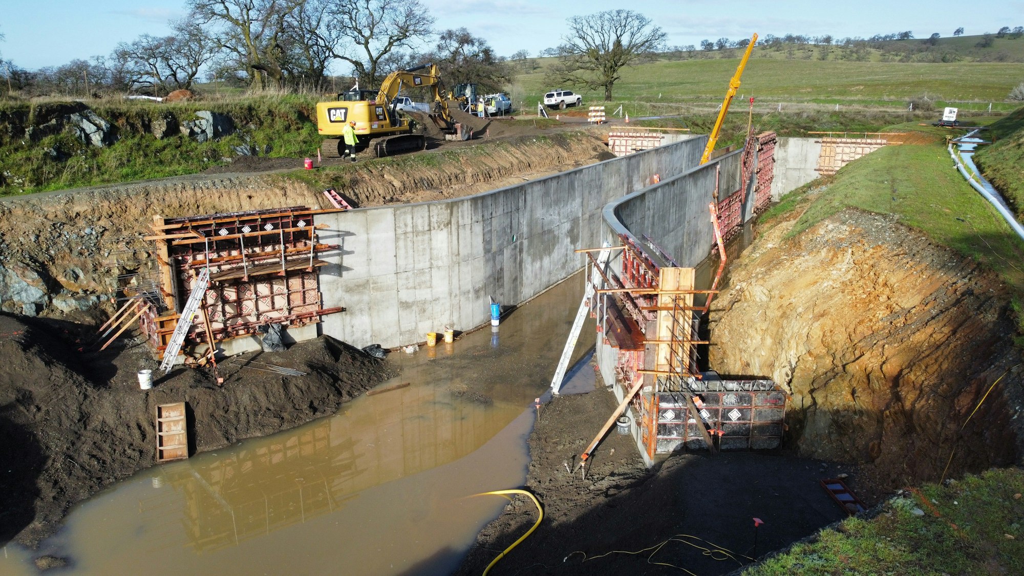Construction site with heavy machinery, concrete walls, scaffolding, and muddy water in a rural landscape.