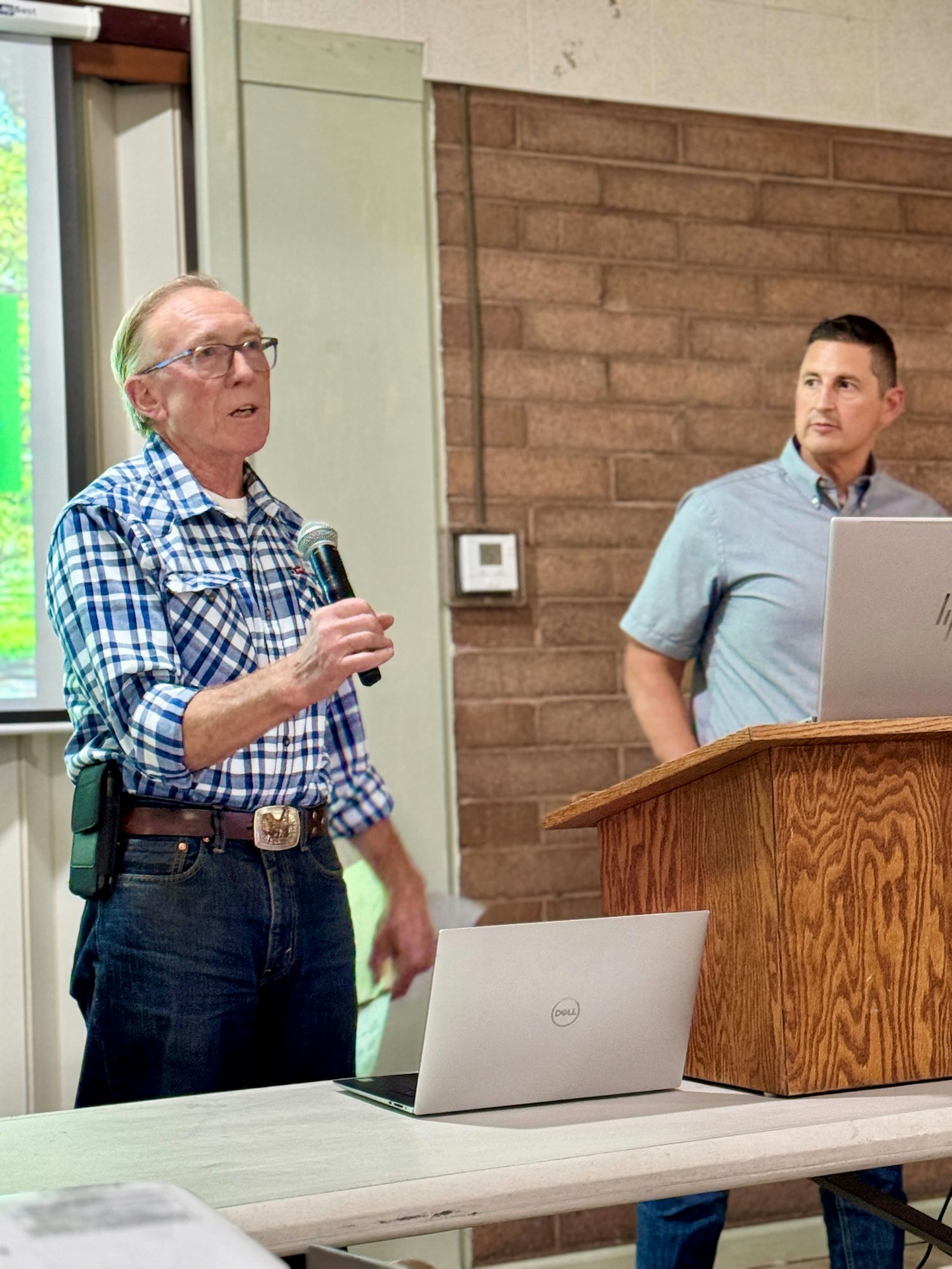 Two men are at a presentation; one is speaking into a microphone, and the other stands near a laptop on a podium.