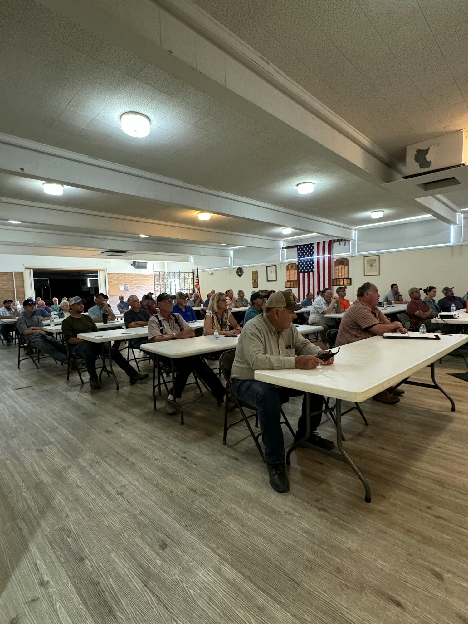 A group of people sitting at tables in a hall, possibly during a meeting or community event, with an American flag in the background.