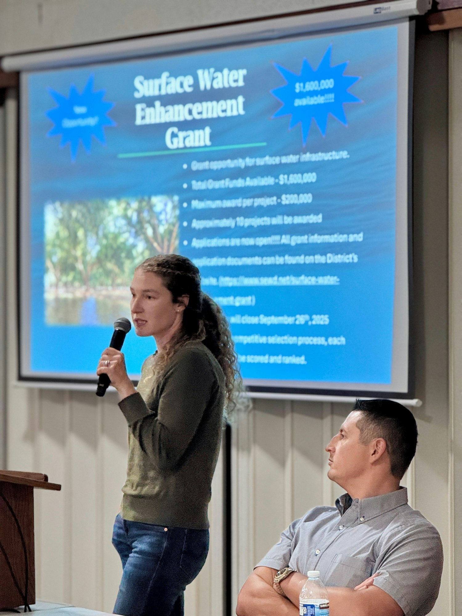 A woman presents a Surface Water Enhancement Grant on a projector screen; a man listens.