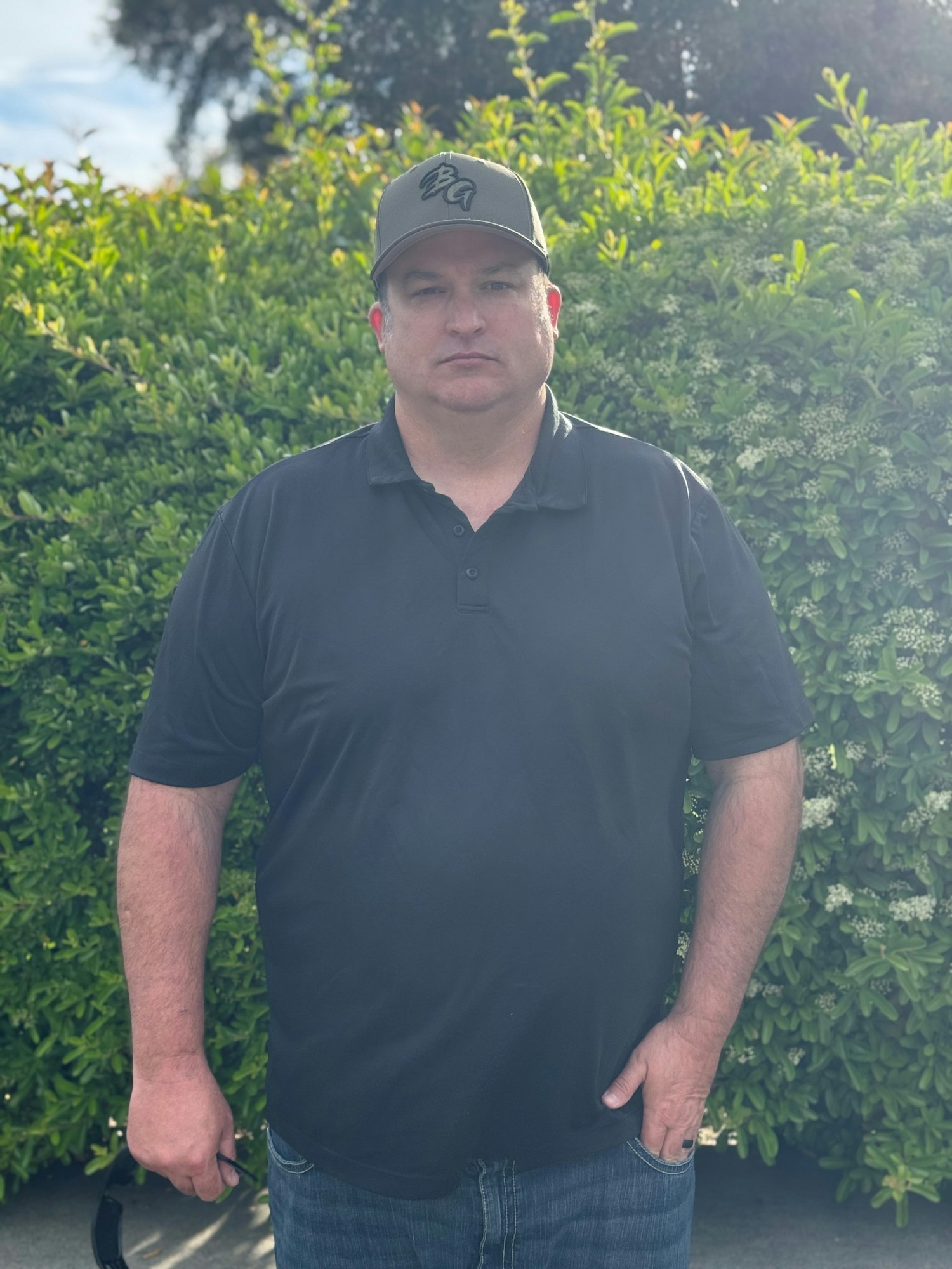 A man in a black polo shirt and a green cap stands in front of a hedge, appearing serious and relaxed.