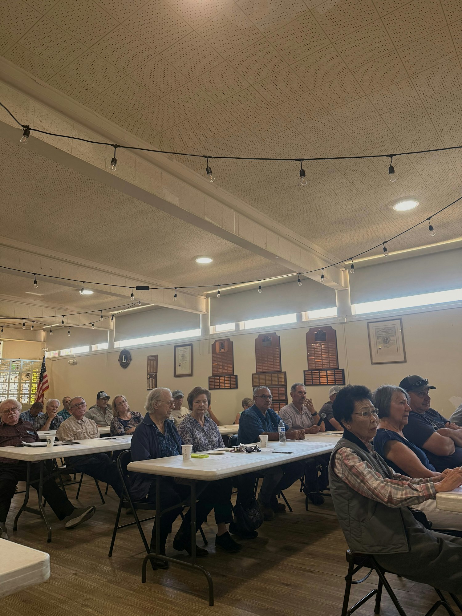 A group of people, mostly older adults, sitting at tables in a well-lit room, possibly attending a meeting or presentation.