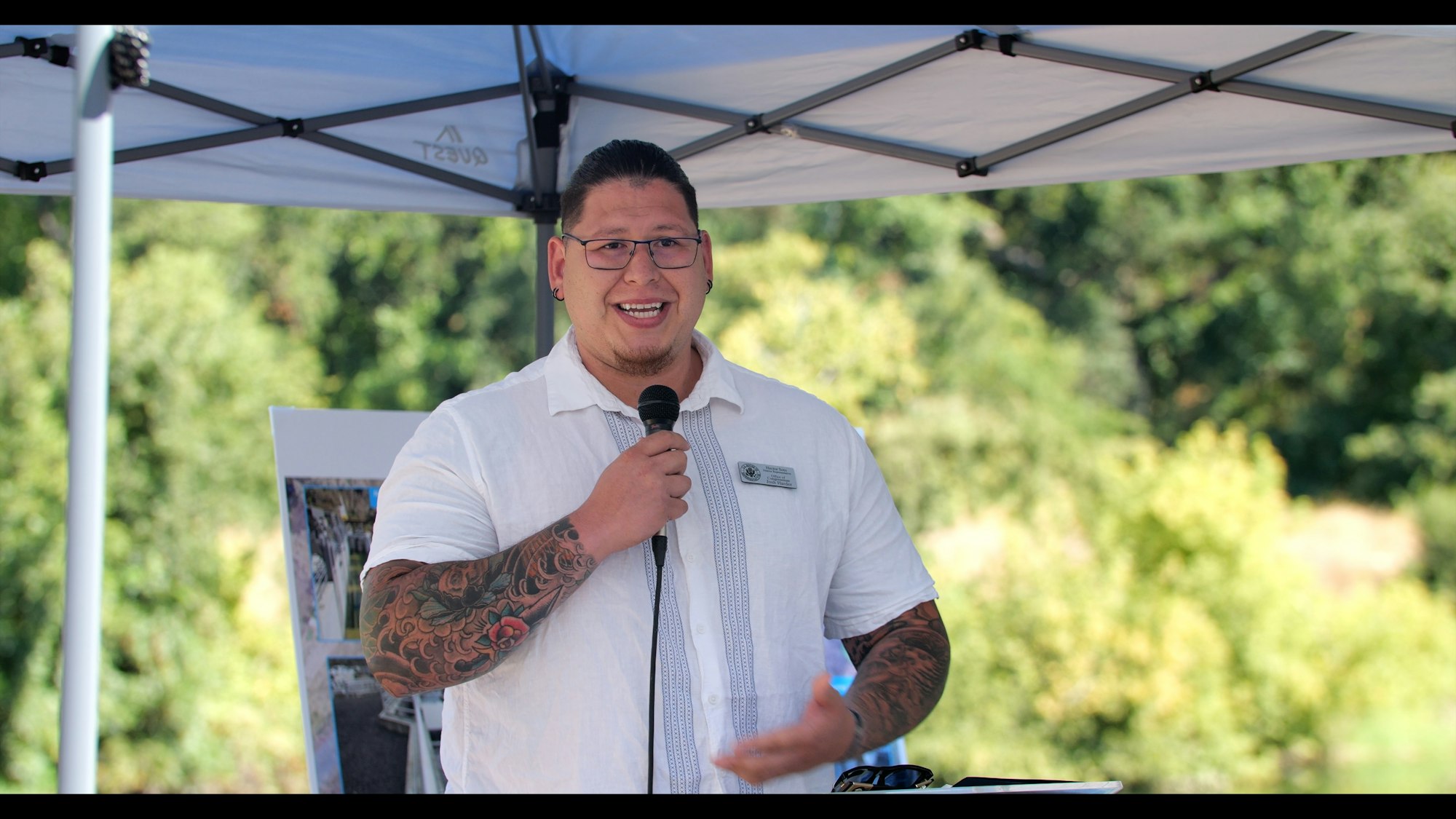 A person speaking into a mic under a canopy, with trees in the background.