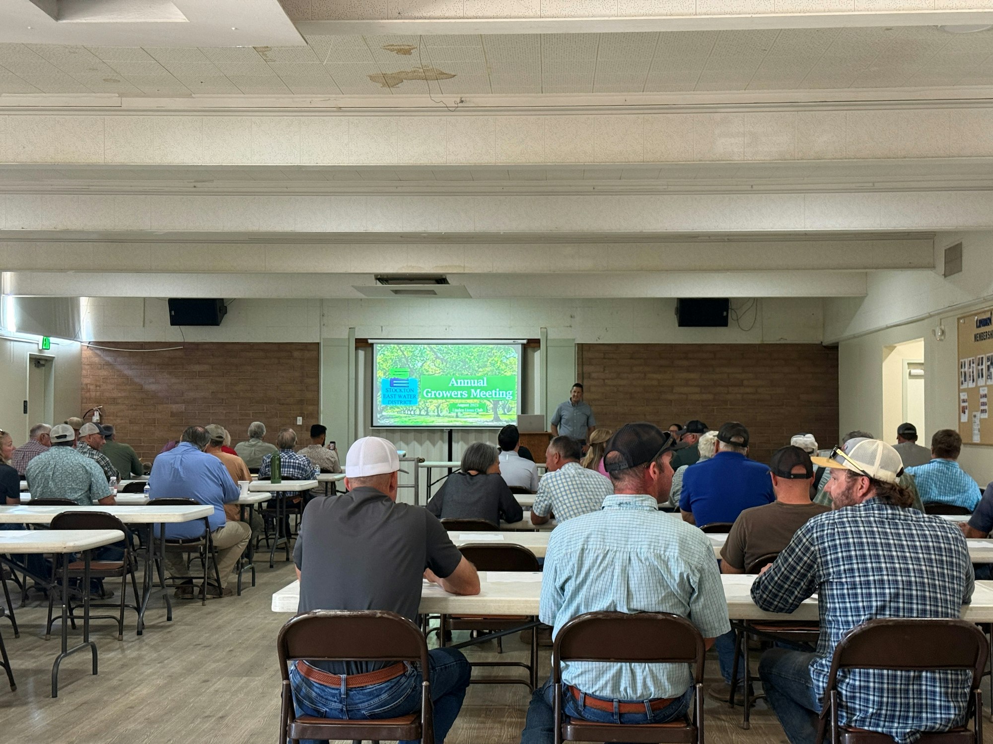 A group of people sit in a room with a presentation titled "Annual Growers Meeting" displayed on a screen in front.