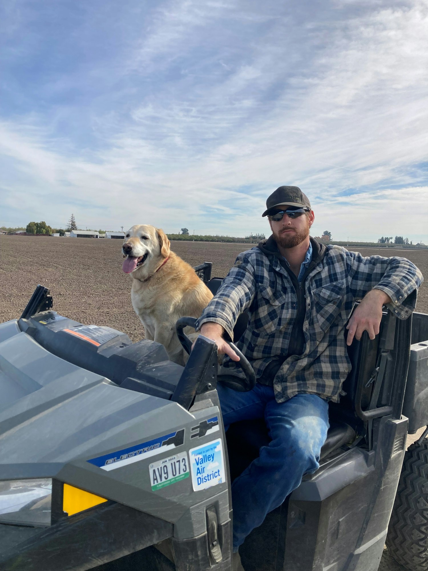 A man in a plaid shirt sits on a vehicle, smiling with a golden retriever beside him against an outdoor backdrop.