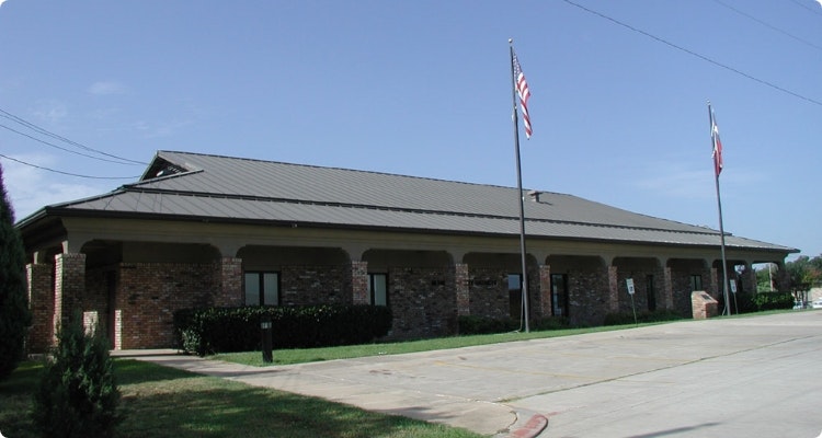A brick building with a metal roof, two flagpoles, and a driveway in front.