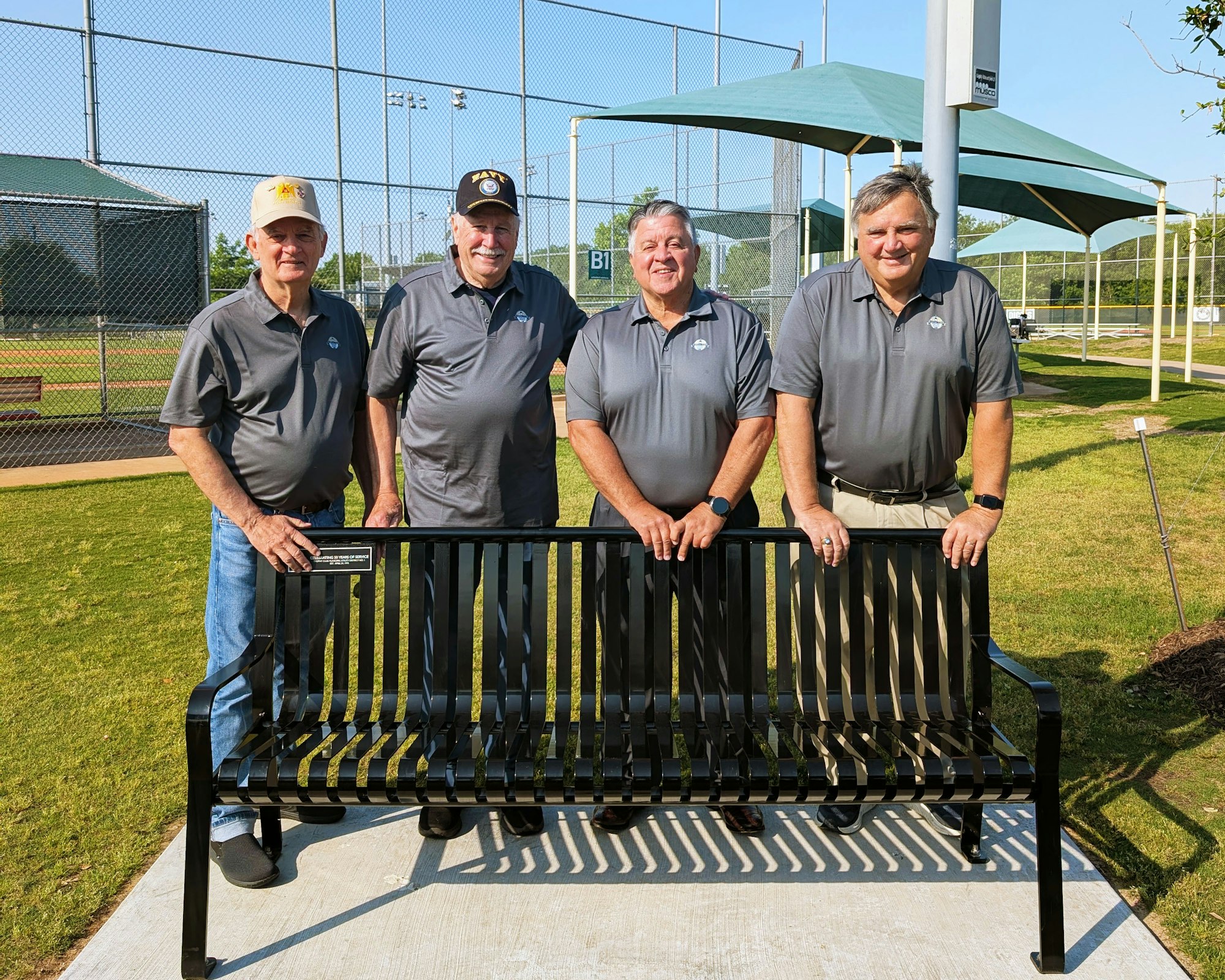 Four men in matching gray shirts standing behind a black bench near a baseball field.