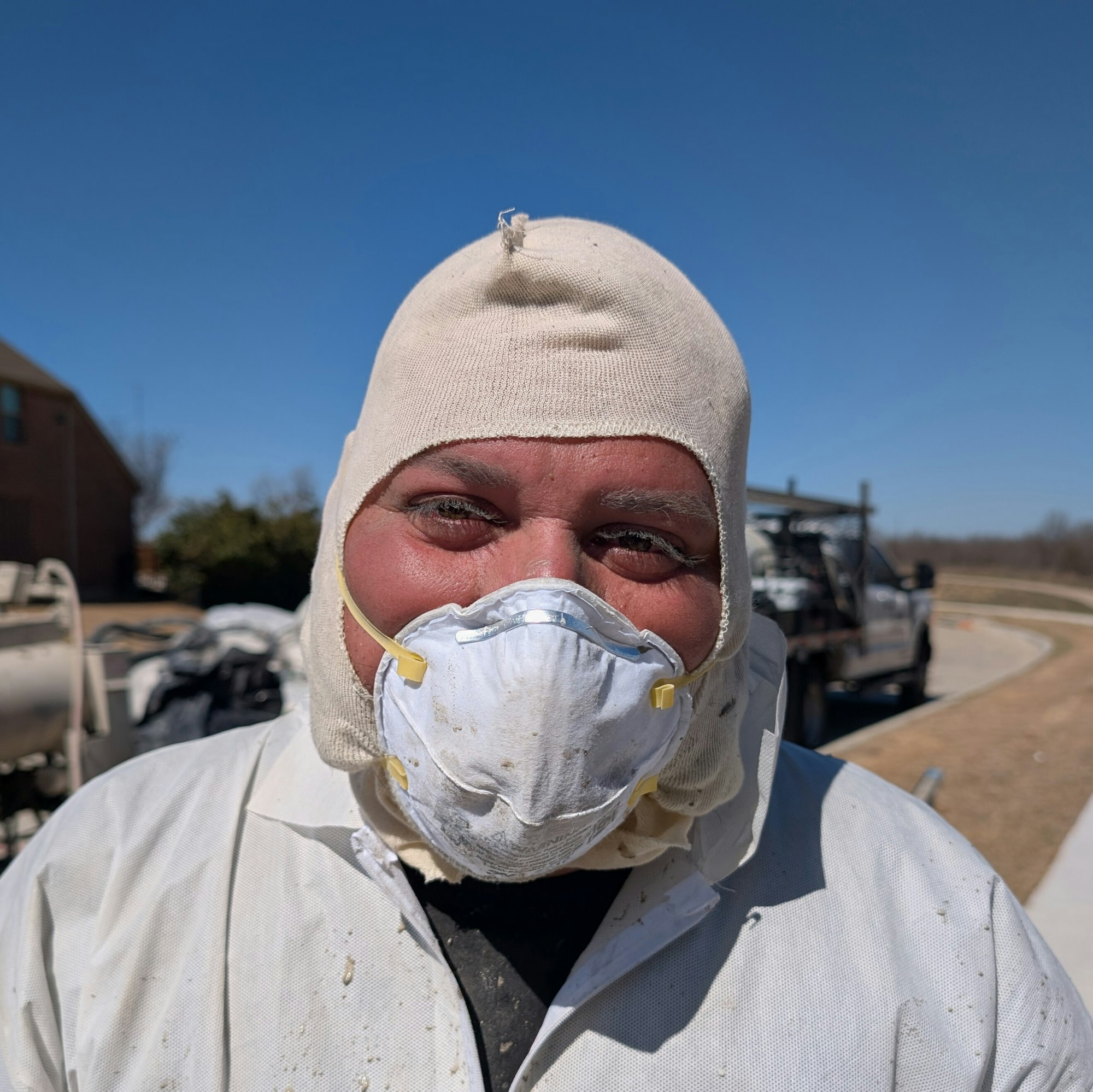 A person in protective clothing and a mask smiles with remnants of sprayed mortar on his eyelashes and eyebrows.