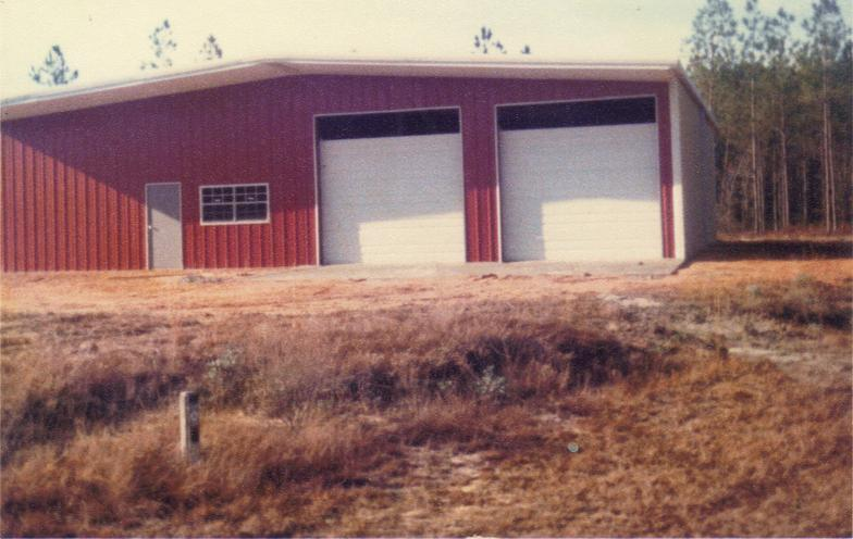 A red warehouse with two large white garage doors, surrounded by grassland and some trees in the background.
