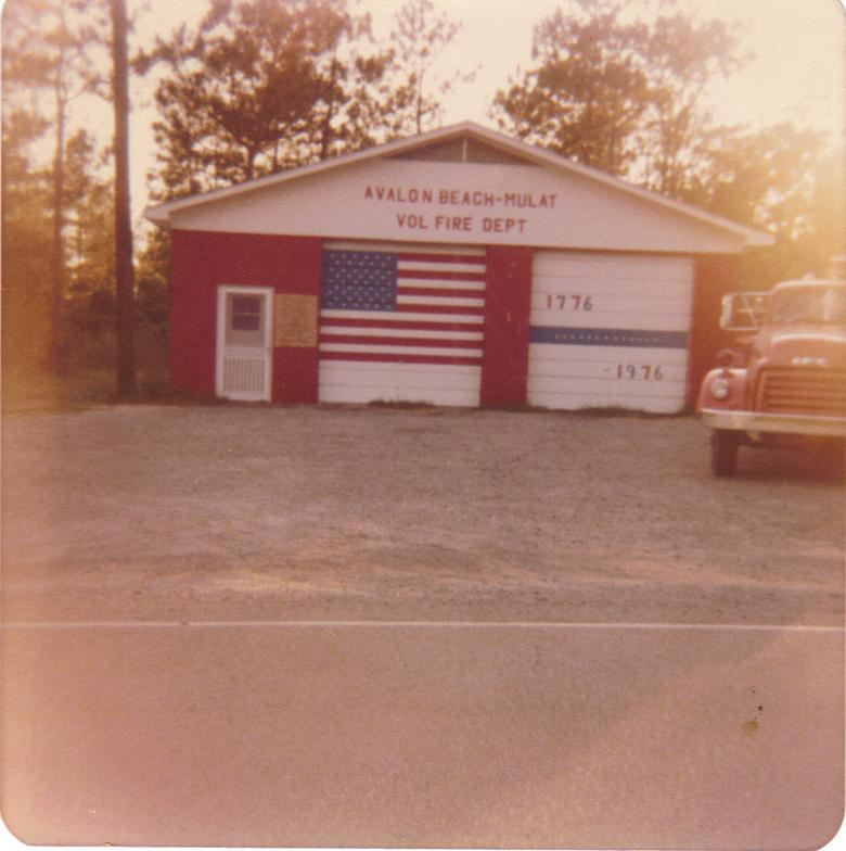 A vintage photo of a small fire department building with American flag motif and a fire truck parked outside.