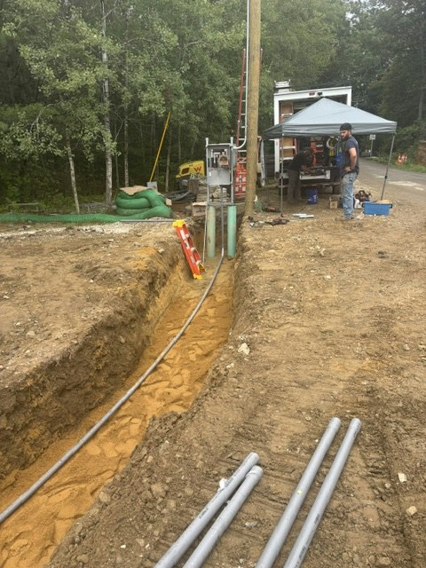 A construction site with a trench, tools, and equipment, likely for laying pipes, surrounded by trees.
