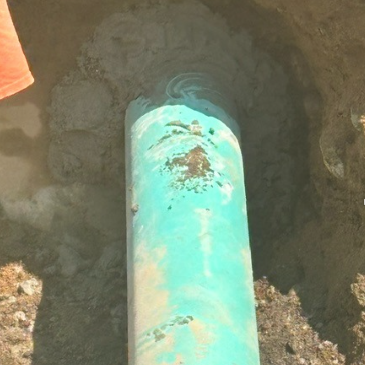 A partially exposed turquoise pipe in a dug-up area, with some surrounding dirt and an orange fabric visible.