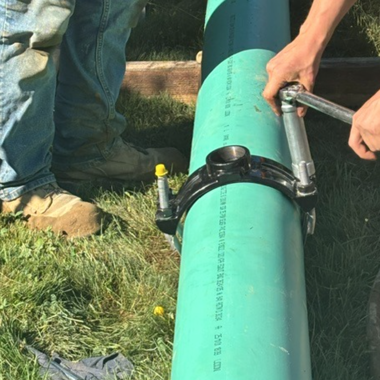 Two construction crew members are are working with a large green pipe, using a socket wrench to secure a fitting along the grassy edge of Donica Road
