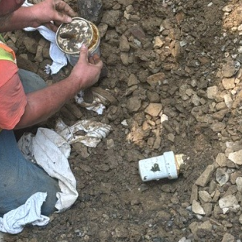A person is digging in soil, holding a canister while another container lies in the ground amidst dirt and rocks.