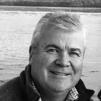 A smiling man in a black and white photo, likely by a body of water, with a casual appearance.