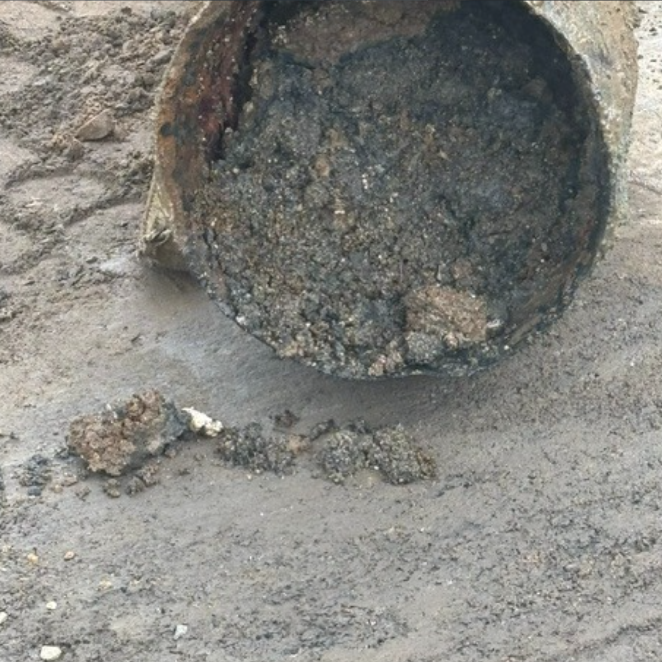 The image shows a rusty, open metal container with thick, dark sludge spilling out onto the ground.