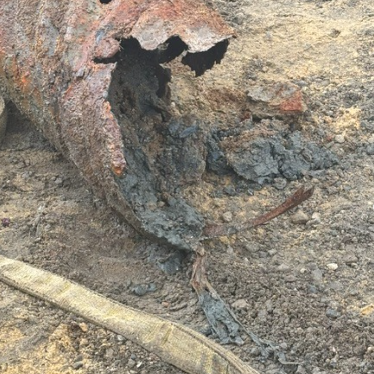 The image shows a rusted, corroded metal object partially buried in dirt, with some debris around it.