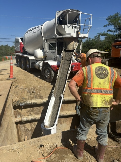 A worker pours concrete from a truck into a construction site, surrounded by equipment and orange traffic cones.