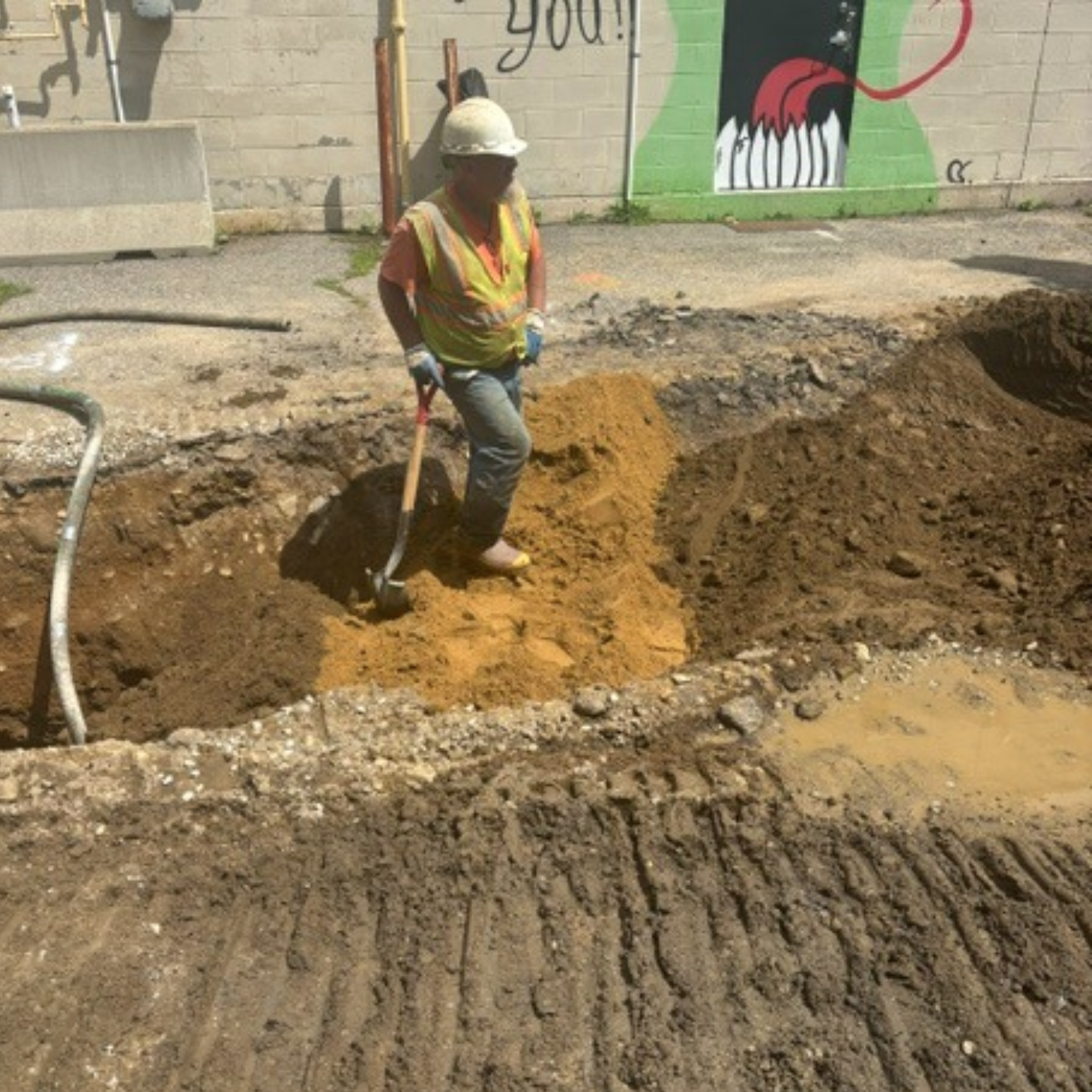 A worker in a yellow hard hat stands in a deep trench, using a shovel to move dirt. A colorful mural is visible in the background.