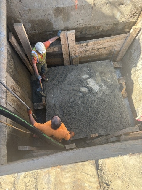 Two construction workers are pouring concrete into a framed area, working on a concrete slab or foundation.