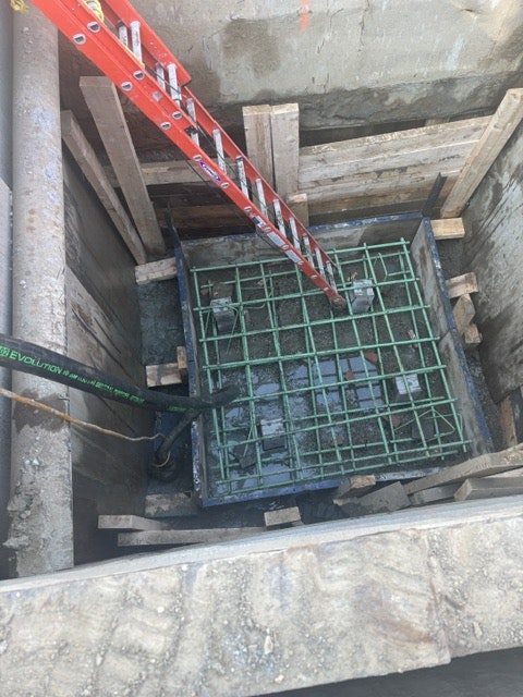 The image shows a construction site with a ladder leading down to a concrete foundation area, featuring a grid structure and wooden supports.