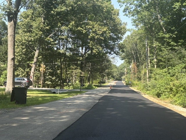 Picture of a tree-lined road with freshly laid asphalt, street lights, and a car parked nearby, indicating road construction or maintenance.
