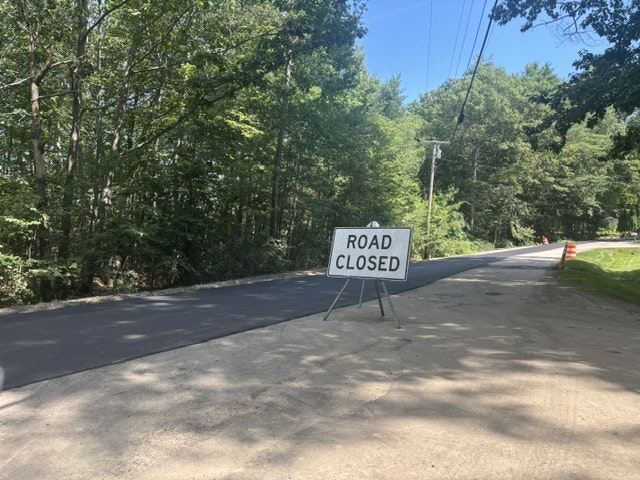 A "Road Closed" sign stands on a paved road surrounded by trees, indicating the road is not accessible.